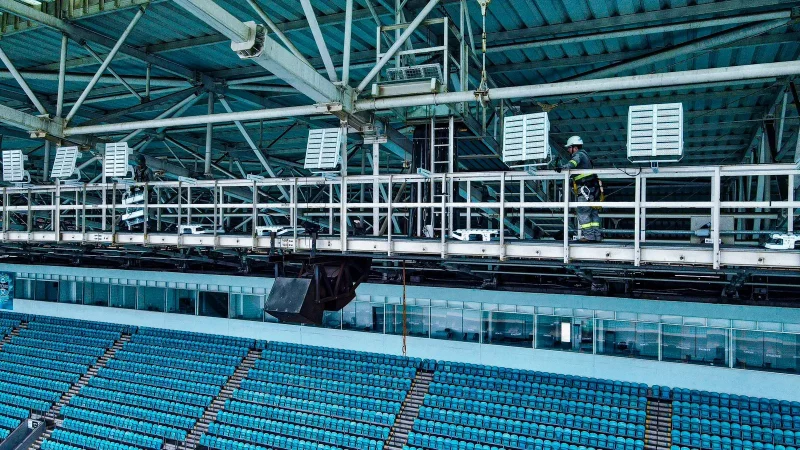 Arena do Grêmio: um funcionário instalador de refletor com capacete na cabeça, no alto das arquibancadas do estádio, trabalhando, ajustando um dos refletores da Arena. 